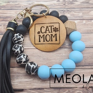 May include: A light blue and black beaded keychain with a wooden charm that says "Cat Mom" and a black tassel. The keychain is made by Meola & Company.