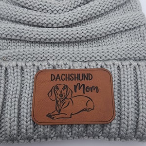 May include: Gray knitted beanie with a faux fur pom pom and a brown leather patch that says "Dachshund Mom" with a dachshund silhouette.