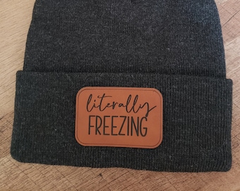 Może przedstawiać: Szary, dzianinowy beret z brązową skórzaną naszywką z napisem "literally freezing". Beret ma puszysty, szary pompon na górze.