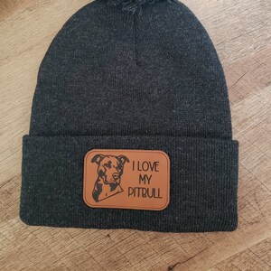 May include: A gray knit beanie with a black pom pom on top. The beanie has a brown leather patch with a black silhouette of a pit bull and the text "I love my pitbull".