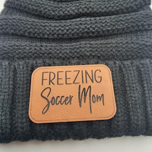Puede incluir: Gorro de punto negro con un parche de cuero marrón que dice "FREEZING Soccer Mom".