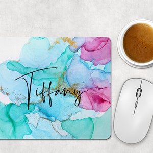 May include: A personalized mousepad with a colorful abstract design in shades of teal, pink, and gold. The name "Tiffany" is written in elegant black script. A white computer mouse and a cup of coffee are next to the mousepad.