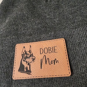 May include: A brown leather patch with a black Doberman Pinscher silhouette and the text "DOBIE Mom" sewn onto a dark gray beanie.