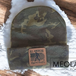 May include: A camouflage beanie with a brown leather patch that says "All American Bigfoot Hunter".