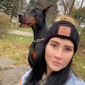 May include: A black knit beanie with a faux fur pom-pom and a brown leather patch featuring a logo. A black and brown Doberman Pinscher dog is in the background, wearing a gold chain collar. The dog is looking to the left.