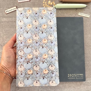 May include: A clear plastic planner cover with a blue and white pumpkin pattern. The cover is placed on top of a black planner with the text "2025" and "COMMON PLANNER" printed on the front.