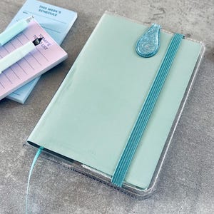 Könnte beinhalten: Ein mintgrüner Planer mit einem türkisfarbenen Gummiband und einem glitzernden türkisfarbenen Lesezeichen. Der Planer befindet sich neben einem Notizblock mit dem Text "This Week's Schedule" und "Today's To Do List". Zwei Stifte sind ebenfalls sichtbar.