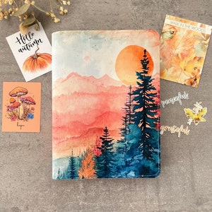 May include: A watercolor painting of a sunset over a mountain range with a forest in the foreground. The sky is a vibrant orange and pink, and the mountains are a soft pink. The trees are a deep blue and green. The words "peaceful" and "grateful" are written in white on the bottom right corner of the image.