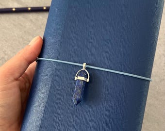 May include: A dark blue journal with a textured cover, secured by a light blue elastic band. A silver pendant with a blue crystal hangs from the band. White flowers and pens are in the background.