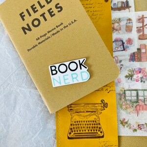 Op de afbeelding: Een bruin Field Notes notitieboekje met een blauwe en witte sticker waarop "BOOK NERD" staat. Het boekje ligt op een geel lederen dagboek met een vintage typemachine-illustratie.