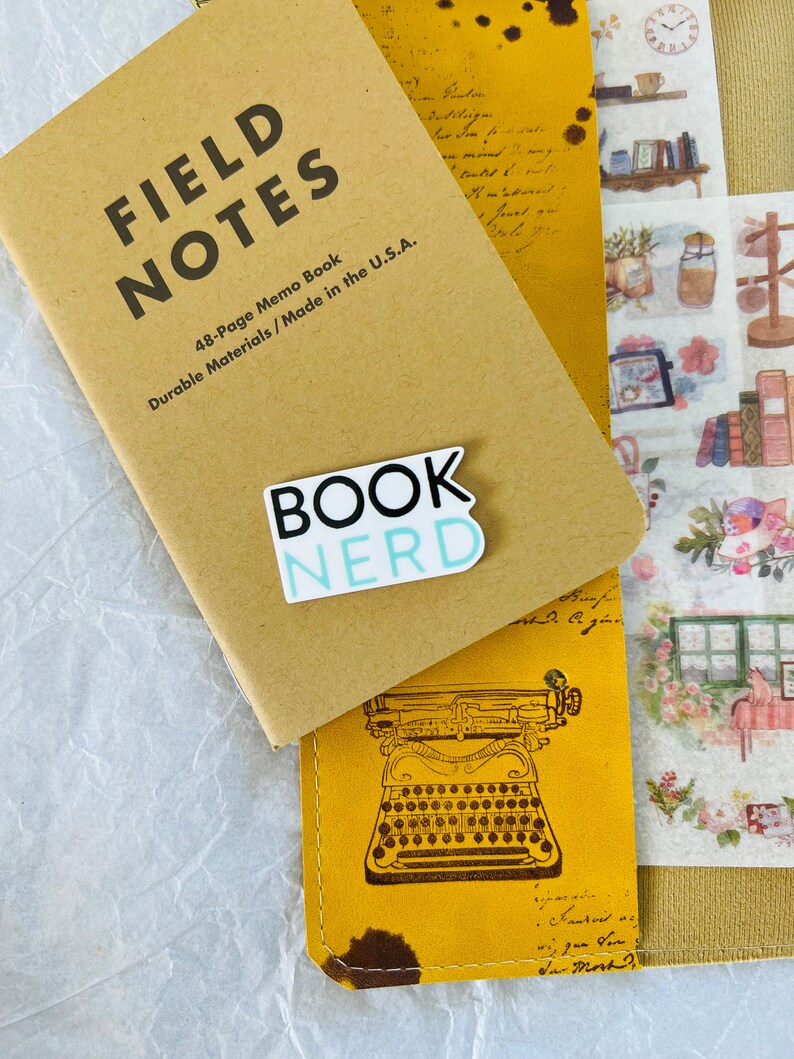 Op de afbeelding: Een bruin Field Notes notitieboekje met een blauwe en witte sticker waarop "BOOK NERD" staat. Het boekje ligt op een geel lederen dagboek met een vintage typemachine-illustratie.