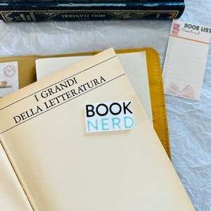 Op de afbeelding: Een close-up van een boek met de titel "I GRANDI DELLA LETTERATURA" en een sticker met de tekst "BOOK NERD". Er is een geel lederen notitieboekje en een roze notitieblok met de titel "BOOK LIST" en de woorden "Books to read" erop.