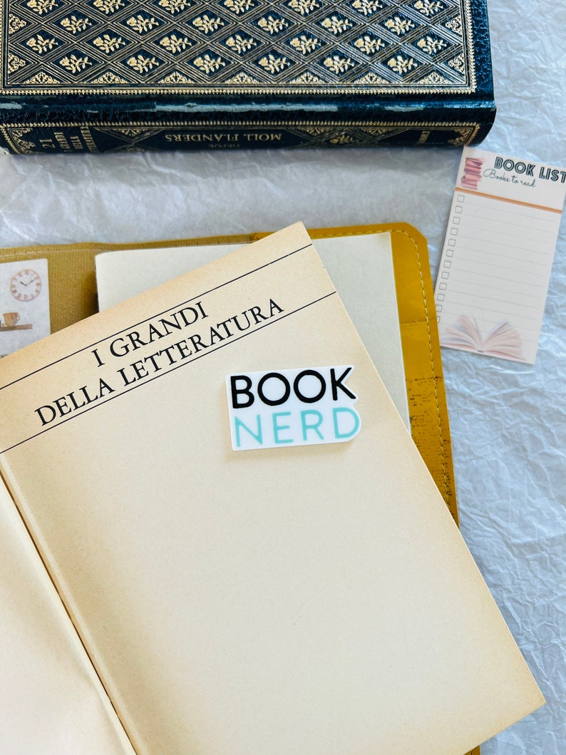 Op de afbeelding: Een close-up van een boek met de titel "I GRANDI DELLA LETTERATURA" en een sticker met de tekst "BOOK NERD". Er is een geel lederen notitieboekje en een roze notitieblok met de titel "BOOK LIST" en de woorden "Books to read" erop.