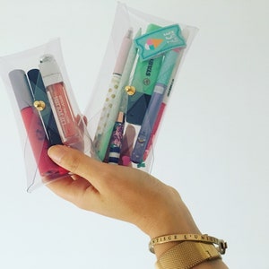 May include: Two clear plastic pouches, one containing lip gloss and lipstick, the other containing pens, highlighters, and a small eraser. The pouches are held in a hand wearing a gold bracelet with the inscription "ANTISCE L'UNICITE".