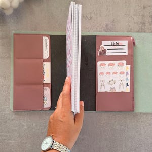 May include: A dusty rose faux leather notebook cover with multiple interior pockets and slots. The cover holds a grid-lined notebook insert and contains various decorative stickers, including one labeled TO DO and another BE AUTHENTIC.