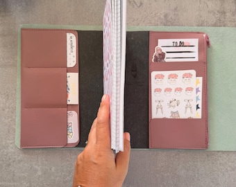 May include: A dusty rose faux leather notebook cover with multiple interior pockets and slots. The cover holds a grid-lined notebook insert and contains various decorative stickers, including one labeled TO DO and another BE AUTHENTIC.