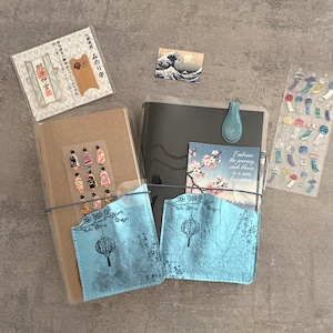 May include: Two notebooks with light blue textured pockets featuring black lantern and branch illustrations. One notebook has a brown cover, the other black. Includes a small Great Wave print, a Meiji Jingu charm, and sticker sheets with kimono and wind chime motifs.