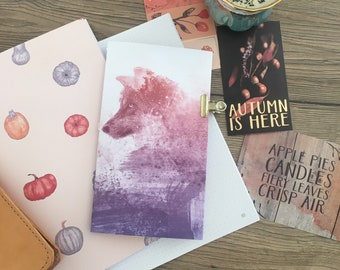 May include: A watercolour illustration of a wolf with a purple and pink gradient background. The illustration is on a white card with a gold clip. The card is on a wooden table with other autumn-themed decorations, including a cup with a candle, a card with the text "Autumn is here", and a card with the text "Apple pies, candles, fiery leaves, crisp air".