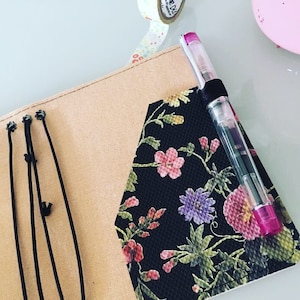 K&ouml;nnte beinhalten: Ein Notizbuch mit schwarzem floralem Muster, mit rosa, lila und roten Blumen. Das Notizbuch ist ge&ouml;ffnet und zeigt ein beiges Innere und schwarze elastische Verschl&uuml;sse. Ein Stift mit rosa Kappe ist in einer Schlaufe auf dem Einband befestigt.