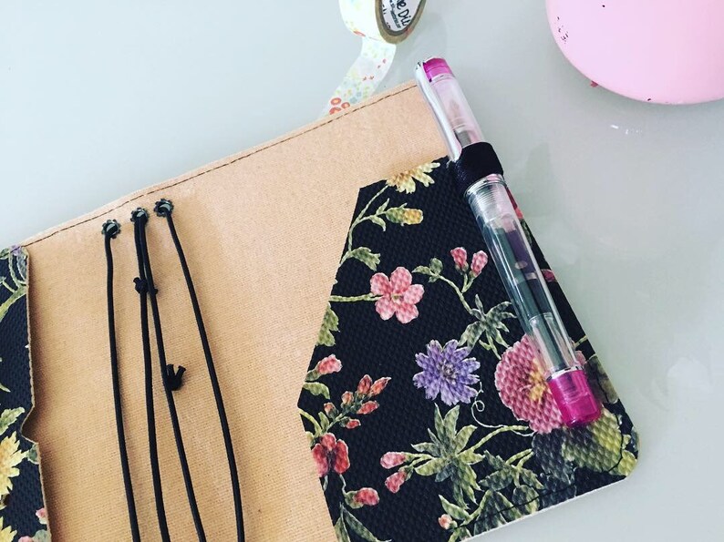 K&ouml;nnte beinhalten: Ein Notizbuch mit schwarzem floralem Muster, mit rosa, lila und roten Blumen. Das Notizbuch ist ge&ouml;ffnet und zeigt ein beiges Innere und schwarze elastische Verschl&uuml;sse. Ein Stift mit rosa Kappe ist in einer Schlaufe auf dem Einband befestigt.