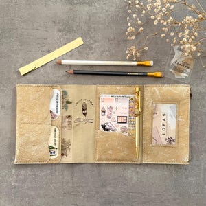Puede incluir: Un organizador de cuero dorado de tres pliegues con múltiples bolsillos interiores, un portalápices con un bolígrafo dorado y ranuras transparentes para tarjetas. El interior tiene un icono de pluma y el texto Made in Italy Crea Tina. Contiene pegatinas y tarjetas con las etiquetas Ideas, Own Your Dreams y Focus on the Good.