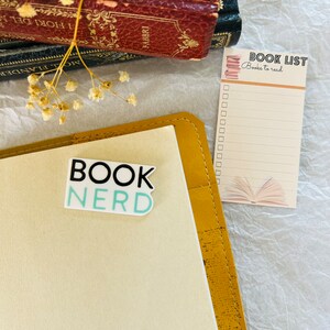 Op de afbeelding: Een geel lederen gebonden dagboek met een sticker waarop "Book Nerd" staat op de voorkant. Een boekenlijst met de tekst "Books to read" ligt op een wit oppervlak naast het dagboek.