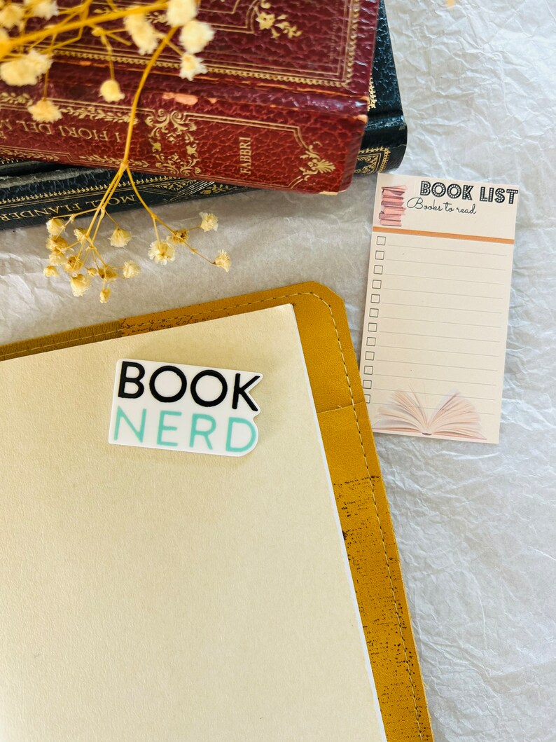 Op de afbeelding: Een geel lederen gebonden dagboek met een sticker waarop "Book Nerd" staat op de voorkant. Een boekenlijst met de tekst "Books to read" ligt op een wit oppervlak naast het dagboek.