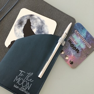 K&ouml;nnte beinhalten: Ein dunkelblaues Stiftetui mit einem silbernen Stift und dem Text "To the Moon". Zwei Untersetzer sind sichtbar: einer mit einem Wolf, der den Mond anheult, und ein weiterer mit dem Text "Manifesting with the Moon". Die Artikel befinden sich auf einer grauen Oberfl&auml;che.
