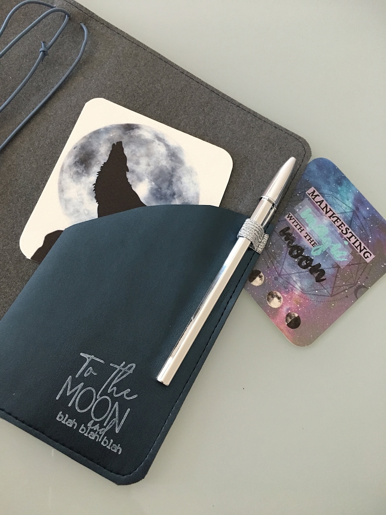 K&ouml;nnte beinhalten: Ein dunkelblaues Stiftetui mit einem silbernen Stift und dem Text "To the Moon". Zwei Untersetzer sind sichtbar: einer mit einem Wolf, der den Mond anheult, und ein weiterer mit dem Text "Manifesting with the Moon". Die Artikel befinden sich auf einer grauen Oberfl&auml;che.