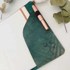 K&ouml;nnte beinhalten: Ein teal-farbener Leder-Notizbuch-Einband mit einem kupferfarbenen Stift in der Tasche. Der Einband hat ein Baumwollblumen-Design und enth&auml;lt ein Notizbuch mit cremefarbenen Seiten. Eukalyptus und Baumwollkapseln sind im Hintergrund.