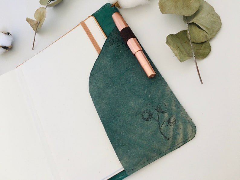 K&ouml;nnte beinhalten: Ein teal-farbener Leder-Notizbuch-Einband mit einem kupferfarbenen Stift in der Tasche. Der Einband hat ein Baumwollblumen-Design und enth&auml;lt ein Notizbuch mit cremefarbenen Seiten. Eukalyptus und Baumwollkapseln sind im Hintergrund.