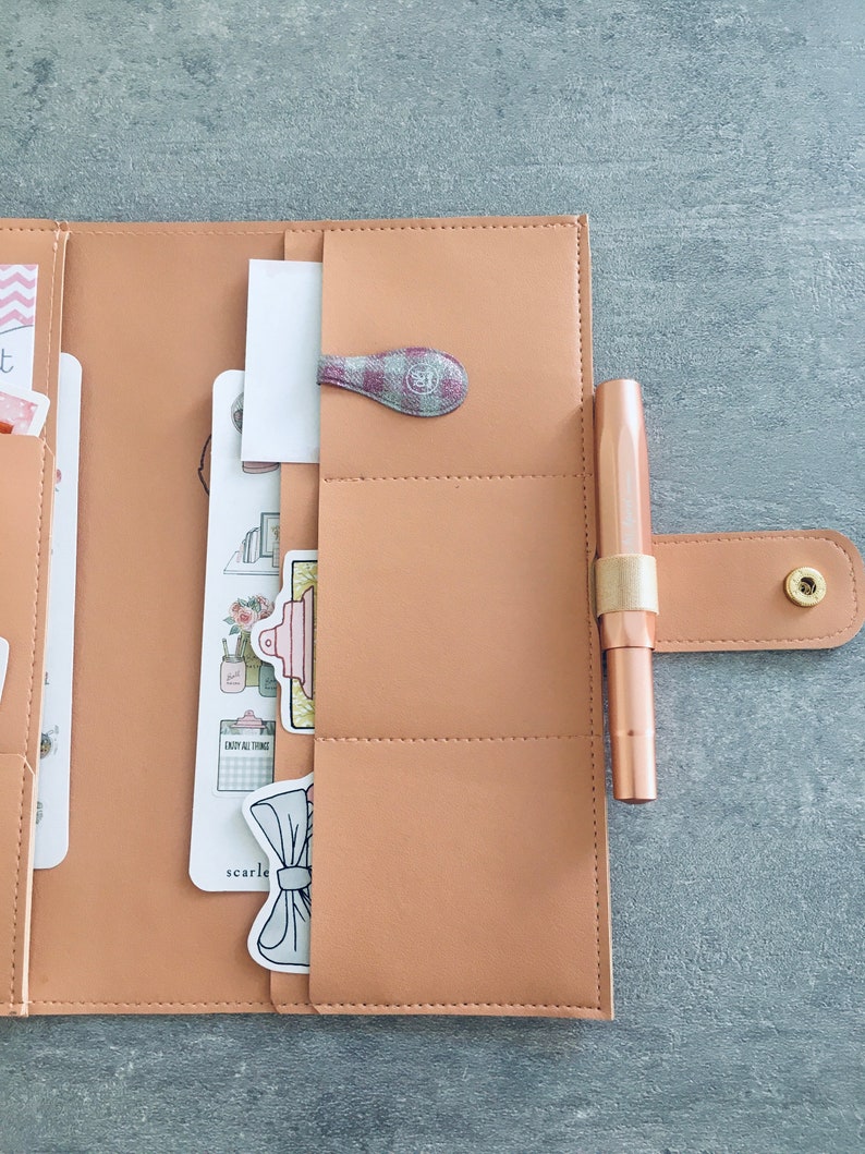 K&ouml;nnte beinhalten: Ein hellbrauner Planer mit einem Stift, einer dekorativen B&uuml;roklammer und mehreren dekorativen Aufklebern. Der Planer ist ge&ouml;ffnet und zeigt Taschen und Eins&auml;tze. Der Stift hat eine metallisch ros&eacute;goldene Farbe. Die B&uuml;roklammer hat ein rosa-graues Karomuster.