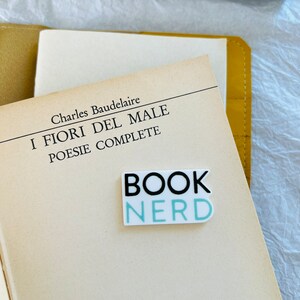 Op de afbeelding: Een witte sticker met zwarte en lichtblauwe tekst die "BOOK NERD" zegt op een boek met de titel "I FIORI DEL MALE" van Charles Baudelaire.
