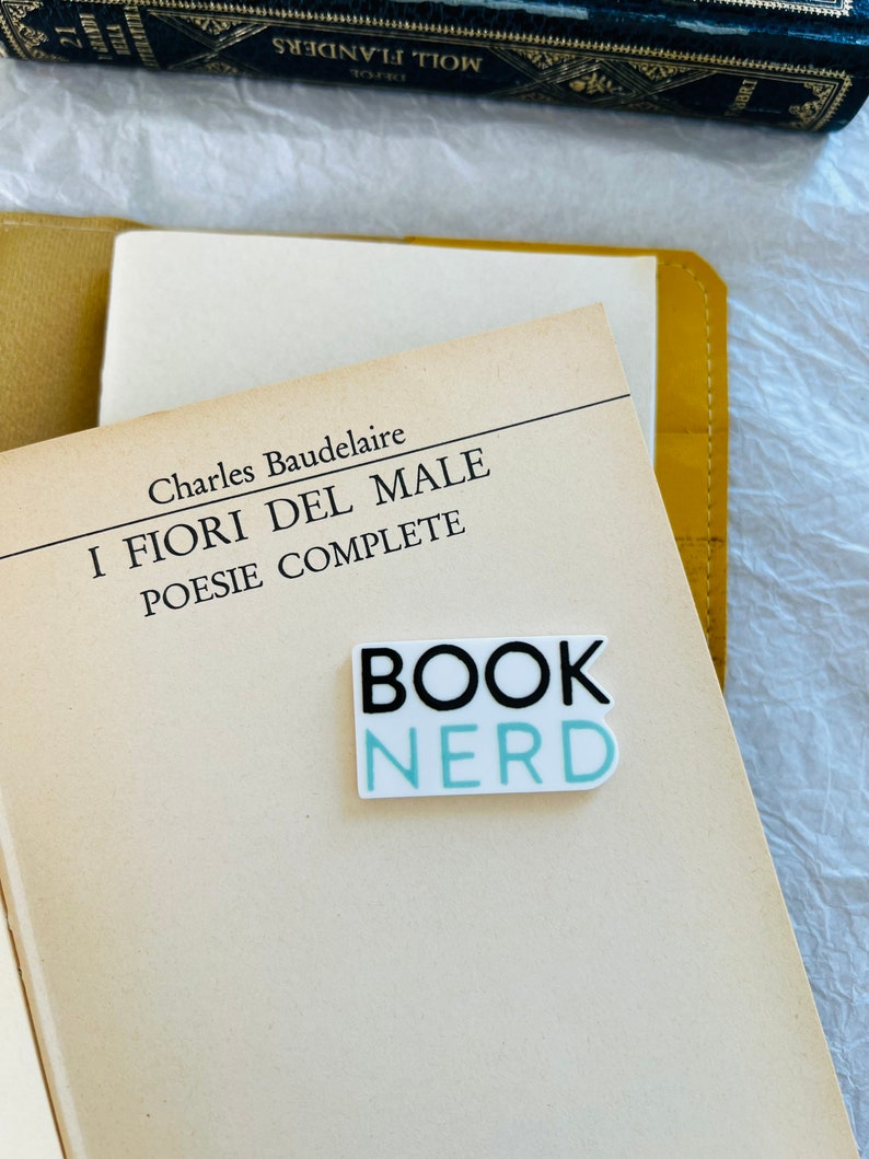 Op de afbeelding: Een witte sticker met zwarte en lichtblauwe tekst die "BOOK NERD" zegt op een boek met de titel "I FIORI DEL MALE" van Charles Baudelaire.