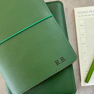 Può includere: Una copertina per planner in pelle verde con le iniziali "R.B." in rilievo sul davanti. La copertina è chiusa e ha un elastico verde avvolto intorno.
