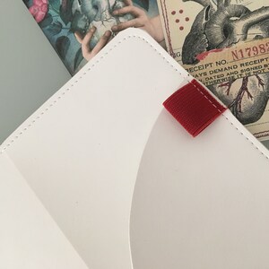 K&ouml;nnte beinhalten: Wei&szlig;er Organizer im Umschlagstil aus Leder mit roter Stofflasche. Der Organizer hat einen gen&auml;hten Rand und ist teilweise ge&ouml;ffnet und zeigt eine bedruckte Karte mit einer Herzillustration und dem Text "RECEIPT NO. N17982".