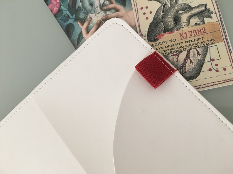 K&ouml;nnte beinhalten: Wei&szlig;er Organizer im Umschlagstil aus Leder mit roter Stofflasche. Der Organizer hat einen gen&auml;hten Rand und ist teilweise ge&ouml;ffnet und zeigt eine bedruckte Karte mit einer Herzillustration und dem Text "RECEIPT NO. N17982".