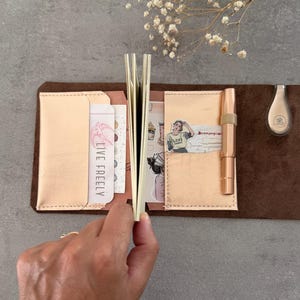 May include: A dark brown leather notebook cover featuring two metallic rose gold interior pockets. The left pocket holds a card with the text LIVE FREELY, while the right pocket contains a rose gold fountain pen and decorative stickers.