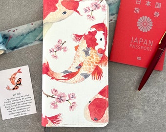 May include: A white leather journal cover with a design of red and orange koi fish swimming through pink cherry blossoms. The text "koi fish" is written in black on a white card with a small illustration of a koi fish. The text "Legend has it that koi scaled a mighty waterfall on the promise of success and happiness at its crest. Rising through the rushing water with grace and determination, they were transformed into golden dragons when they reached the top." is written on the card.