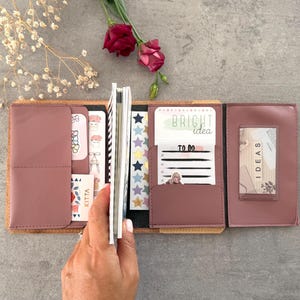 May include: A dusty rose trifold organiser featuring multiple interior pockets and slots. The organiser contains various stationery items, including cards with the text BRIGHT idea, TO DO, and IDEAS, along with sheets of star-shaped stickers.