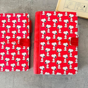 May include: Two red notebooks with a Snoopy pattern. The notebooks have a red leather closure. The Snoopy pattern is white Snoopy figures on a red background. A vintage comic strip is in the background.