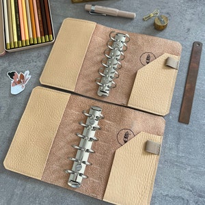 Puede incluir: Dos carpetas de anillas de cuero beige abiertas con herrajes plateados, sobre una superficie gris. También hay una lata de lápices de colores, un bolígrafo y una regla. Las carpetas tienen un bolsillo y un cierre elástico.