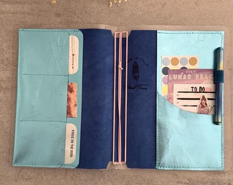 May include: A teal and navy blue leather notebook cover featuring light pink elastic bands, multiple interior pockets, and a pen loop. The cover contains various cards and notes with text including "FOCUS ON THE GOOD", "HIKE LUNAR PEAK", and "TO DO".