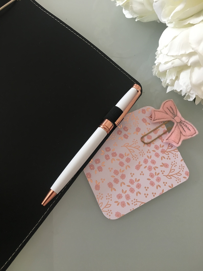 K&ouml;nnte beinhalten: Ein wei&szlig;er Stift mit ros&eacute;goldenen Akzenten liegt auf einem schwarzen Notizbuch. Ein rosa blumig gemustertes Etikett mit einer rosa Schleifen-B&uuml;roklammer befindet sich neben dem Stift. Wei&szlig;e Blumen sind im Hintergrund.