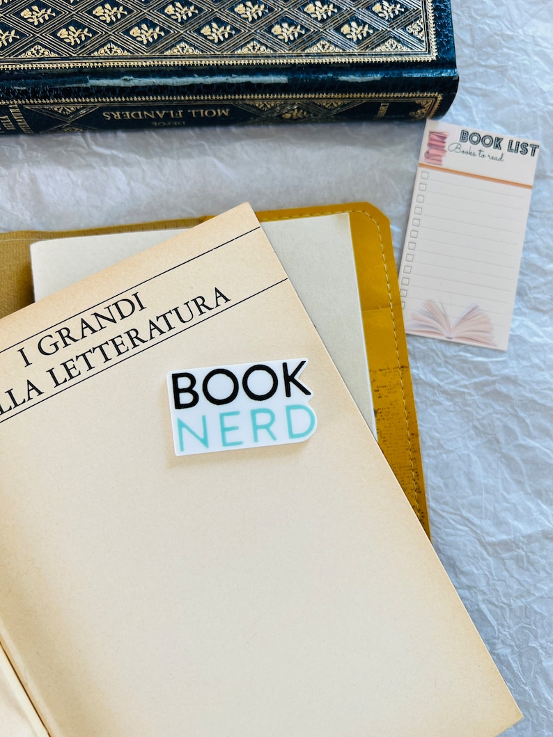 Op de afbeelding: Een close-up van een boek met de titel "I GRANDI DELLA LETTERATURA" en een sticker met de tekst "BOOK NERD" op de kaft. Er is een geel notitieblok met de woorden "BOOK LIST" en "Books to read" erop.