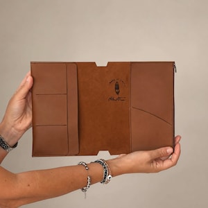 May include: A tan leather document holder featuring a tri-fold design with multiple interior pockets and slots. The centre panel displays a stamped logo with the text Made in Italy and a feather icon. The exterior features a side zipper closure.