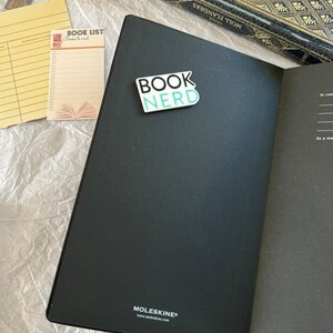 Op de afbeelding: Een zwart Moleskine-notitieblok met een turquoise blauwe sticker waarop "BOOK NERD" staat op de voorzijde. Het notitieblok is open op een lege pagina. Een gele checklist met de woorden "BOOK LIST" en "Books to read" is zichtbaar op de achtergrond.