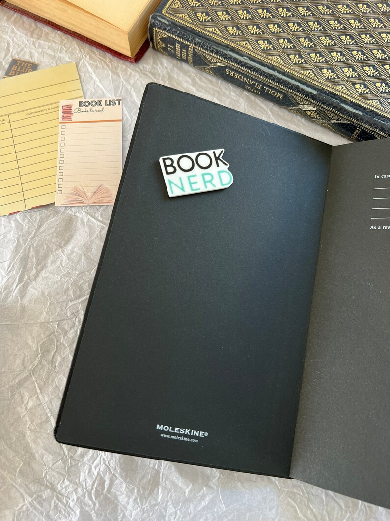Op de afbeelding: Een zwart Moleskine-notitieblok met een turquoise blauwe sticker waarop "BOOK NERD" staat op de voorzijde. Het notitieblok is open op een lege pagina. Een gele checklist met de woorden "BOOK LIST" en "Books to read" is zichtbaar op de achtergrond.