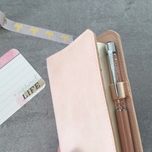 K&ouml;nnte beinhalten: Ein hellrosa Lederjournal mit einem Stift und einer Karte mit der Aufschrift "captured LIFE". Eine Rolle Washi-Tape mit goldenen Schleifenakzenten ist ebenfalls sichtbar. Das Journal hat eine weiche, neutrale Farbe.