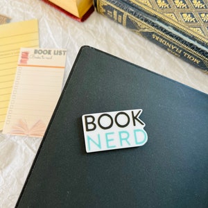 Op de afbeelding: Een zwart notitieboek met een witte sticker waarop "BOOK NERD" in zwart en blauw staat.